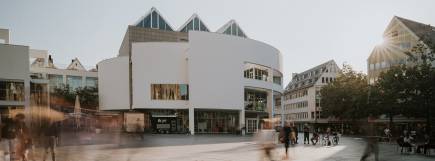 Weitwinkelaufnahme des Stadthauses Ulm mit umliegender urbaner Architektur. Die helle Fassade des Gebäudes hebt sich von der Umgebung ab