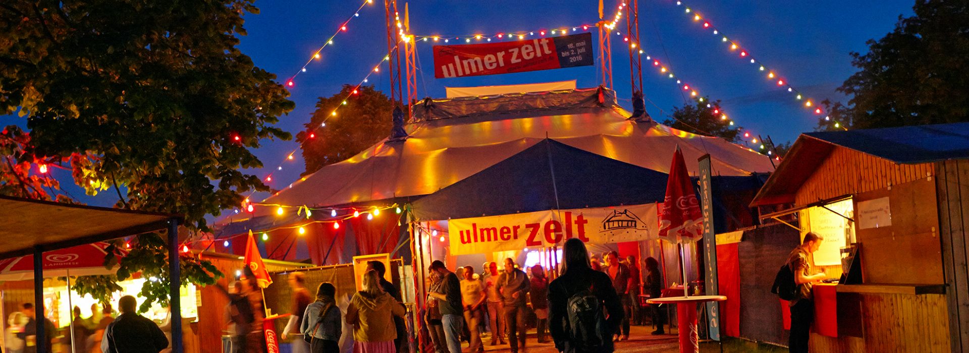 Beleuchtetes Zirkuszelt bei Nacht mit Besuchern und bunten Lichterketten.