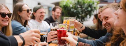 Gruppe junger Menschen sitzt bei Tageslicht an einem Tisch im Freien und unterhält sich lebhaft.