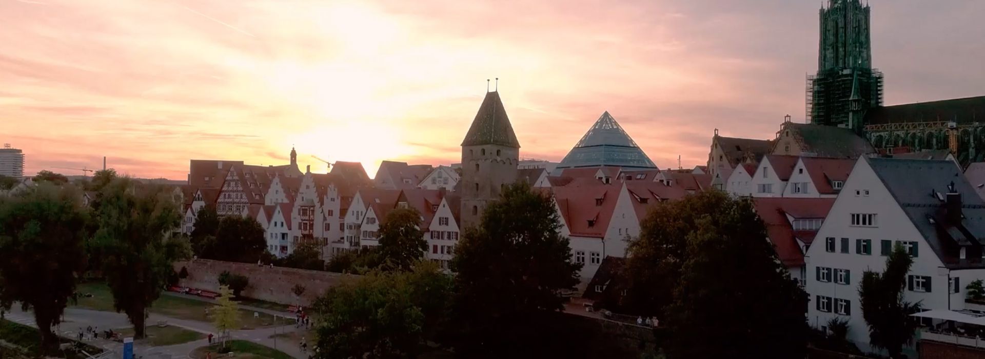 Häuserzeile mit historischem Stadtturm, dem Ulmer Münster und moderner Glaspyramide bei Abenddämmerung in Ulm.