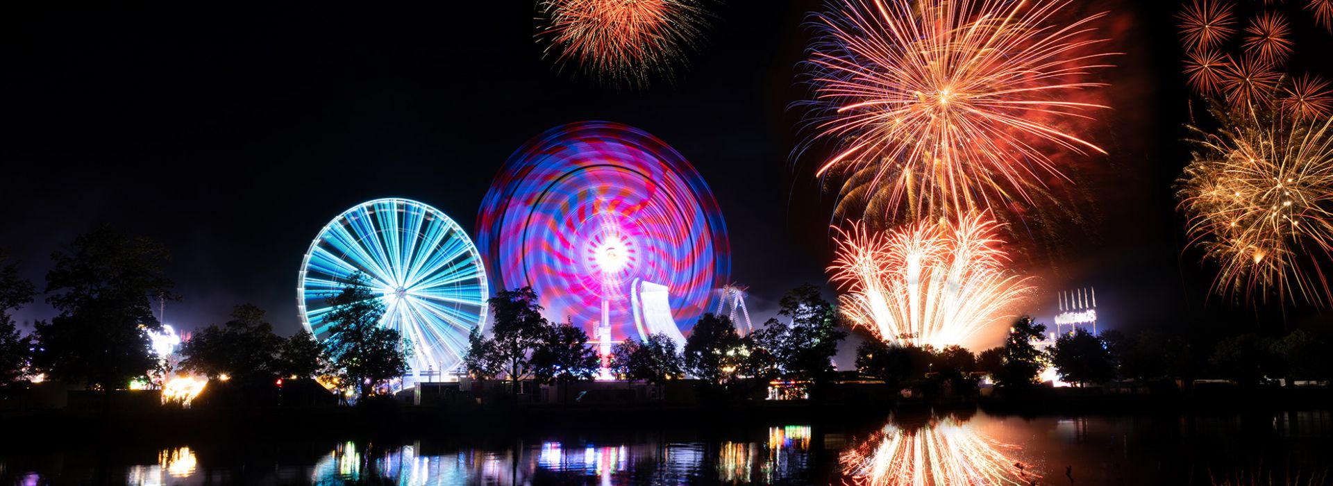 Funkelndes Feuerwerk über beleuchtetem Riesenrad und Volksfest bei Nacht, Spiegelung im Wasser.
